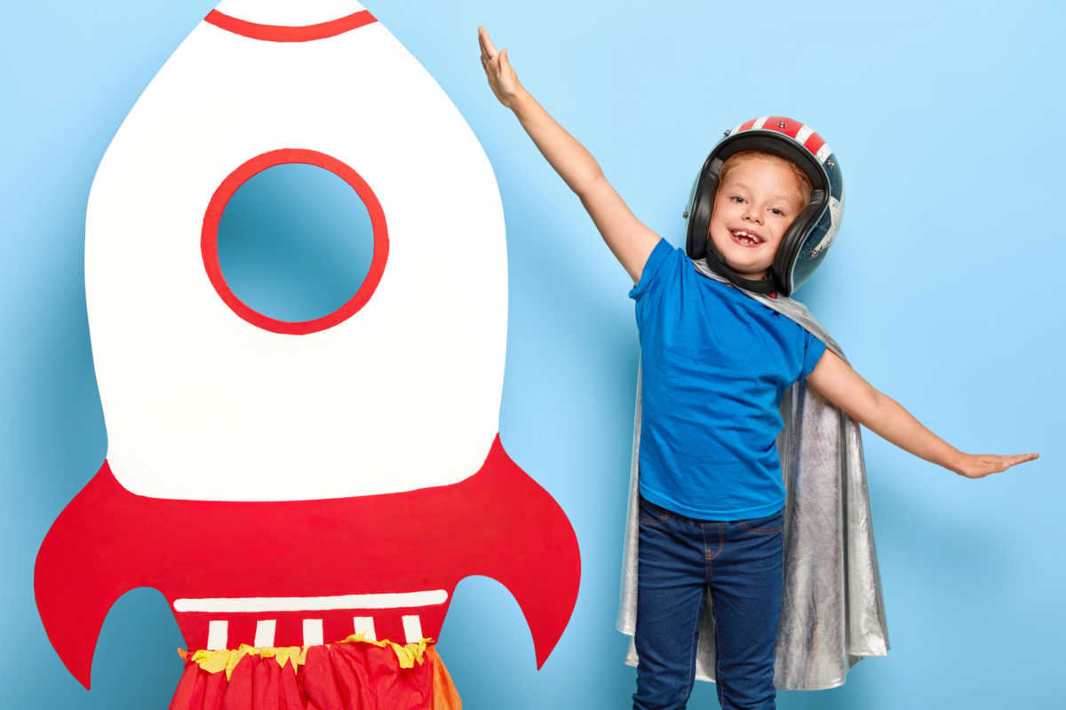 Cute happy female child plays astronaut, wears flying helmet and cape, spreads arms sideways, feels freedom and power, stands near toy rocket, dreams of becoming spaceman. Childhood and fun concept